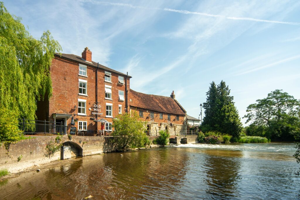 A scenic view of the historic Old Mill by a river in England, surrounded by lush greenery.